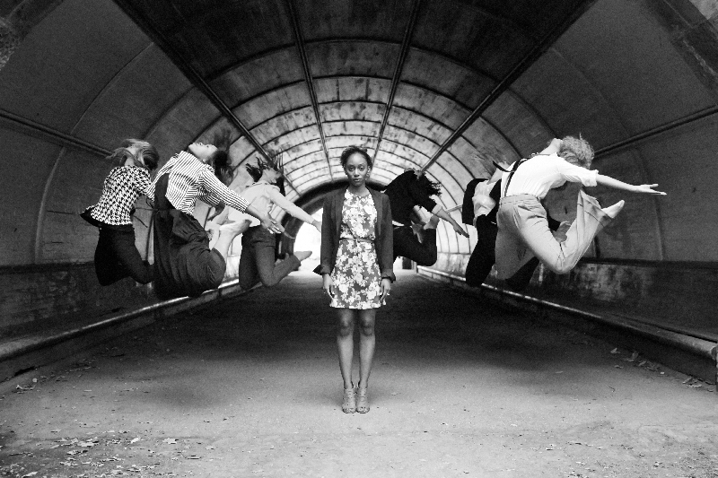 Jamal Jackson Dance Company jump in the air with their chests and heads to the sky while their legs bend and arms sweep behind them. One dancer stands looking straight out in the camera